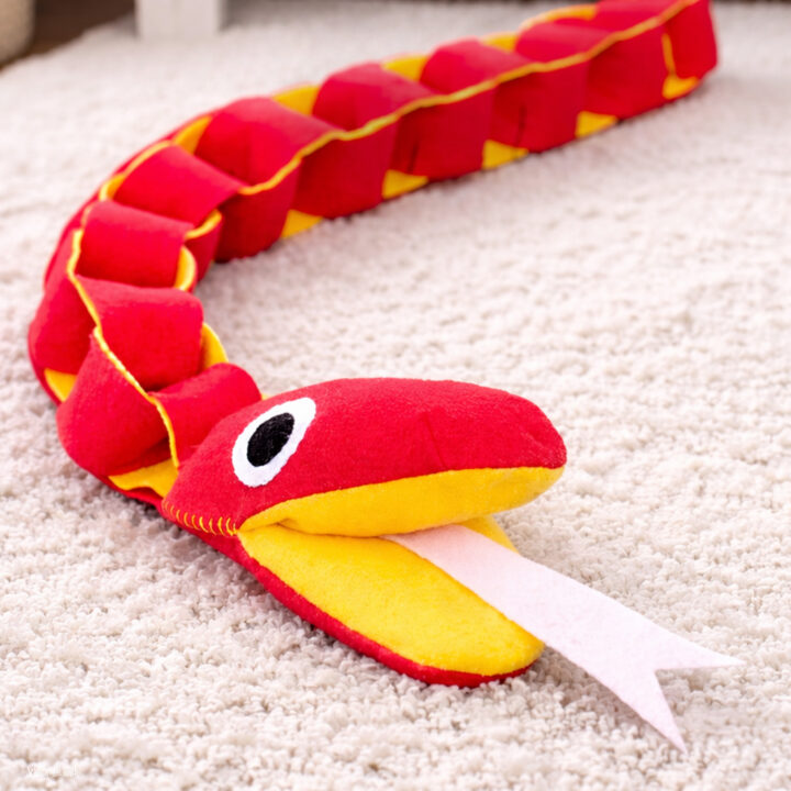Felt chain snake plush sewing pattern listing image with a finished chain snake plush posed in a simple styled setting. Cute handmade stuffed snake from Scratch and Stitch with linked segments and a friendly plush face. Beginner-friendly plush toy sewing pattern for a fun felt sewing project.