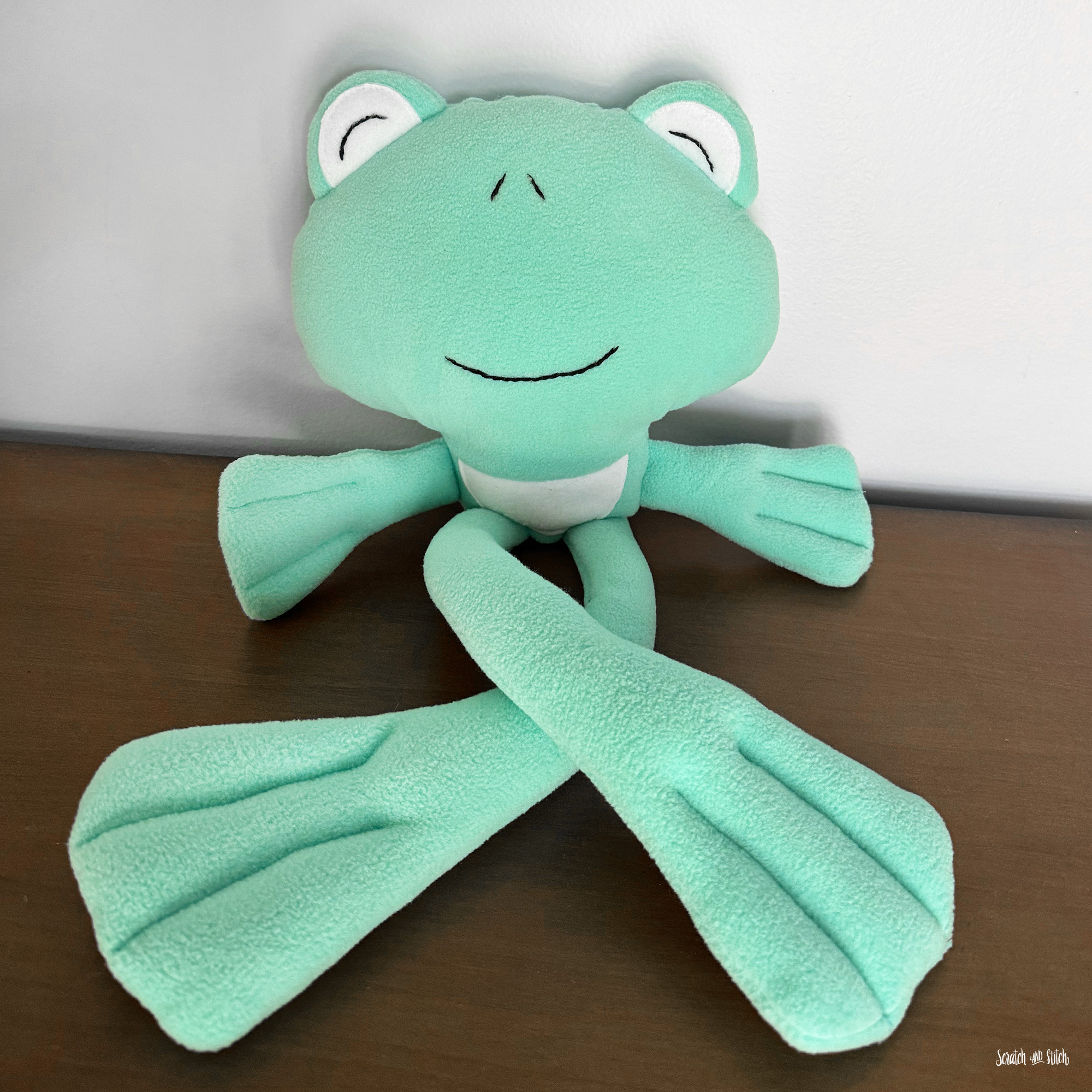 Handmade frog plush sewing pattern sample with long floppy legs and webbed feet, sewn in soft mint fleece and displayed on a dark wood surface by Scratch and Stitch.