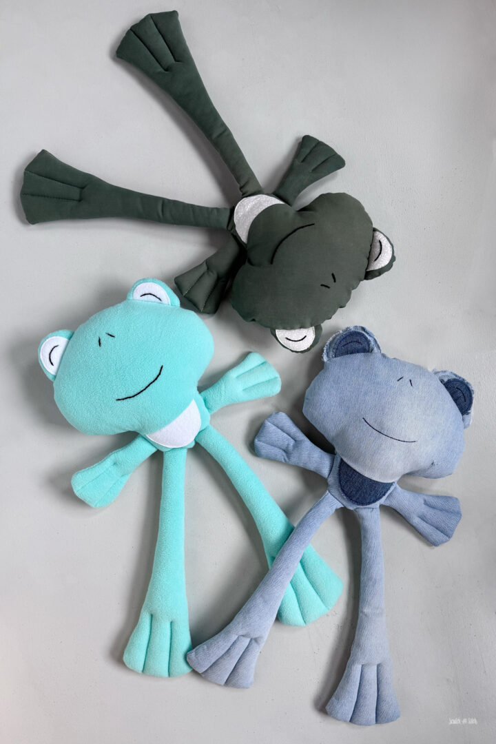 Flat lay photo featuring multiple finished frog plush variations arranged together, showcasing different fabric looks while keeping the same long-legged shape and webbed feet design. Handmade stuffed frogs with embroidered faces and soft eye details. Scratch and Stitch frog plush sewing pattern inspiration image for using different fabrics.