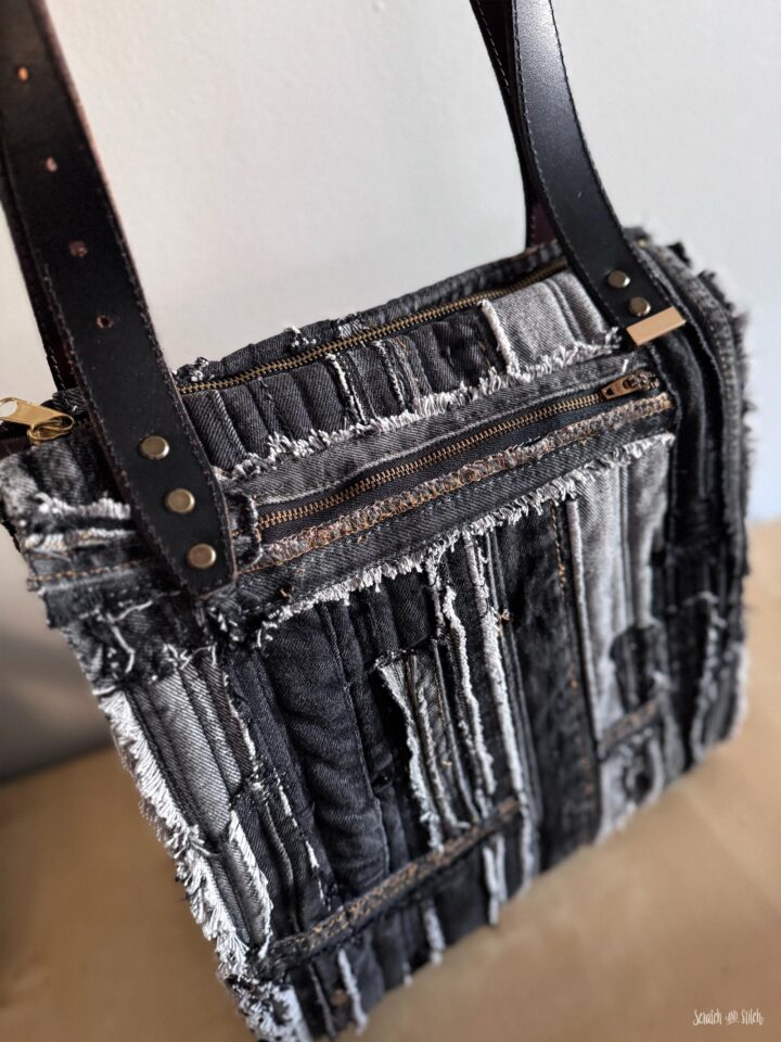 Detail view of an upcycled denim tote bag with an exterior zipper pocket, repurposed leather belt handles, brass rivets, exposed seams, and layered black denim texture.