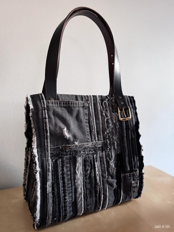 Finished upcycled denim tote bag made from black and gray denim scraps with frayed seams, exterior pocket details, leather belt handles, and a structured handmade bag shape.