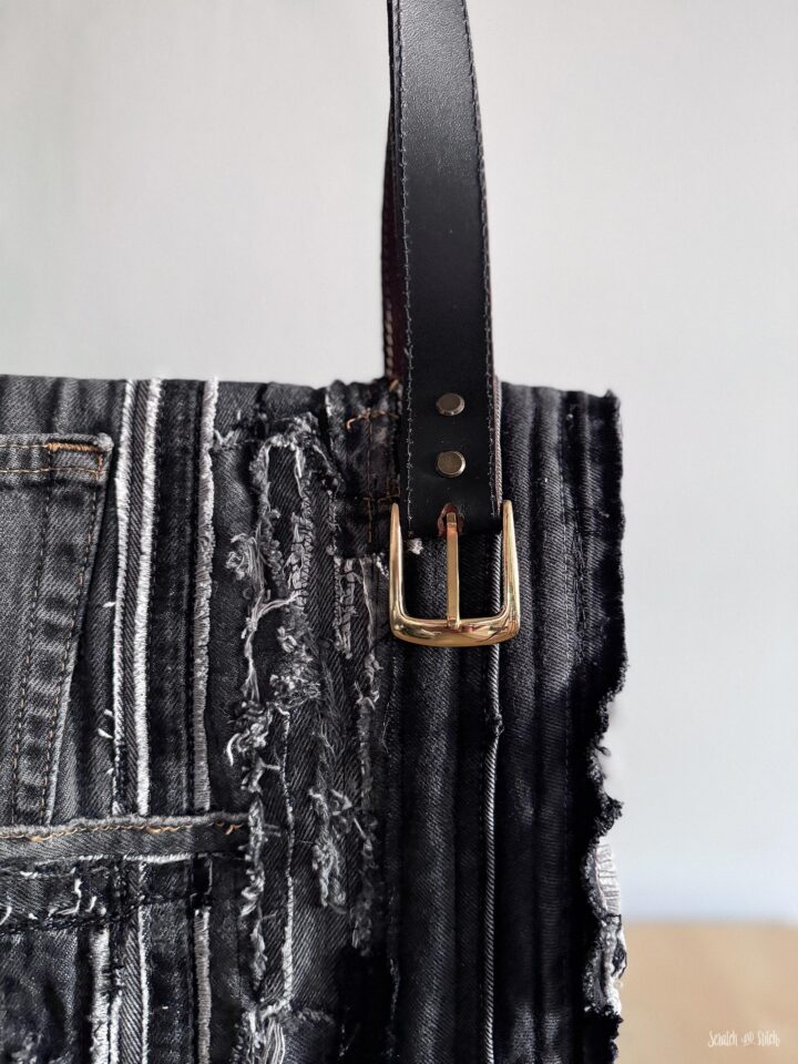 Close-up of an upcycled denim tote bag with a repurposed leather belt handle, brass buckle, rivets, exposed denim seams, and textured black denim patchwork.